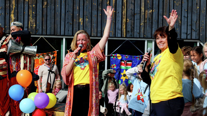 To kvinner i gule T-skjorter holder mikrofoner og vinker på en utendørsfestival med barn og fargerike ballonger.