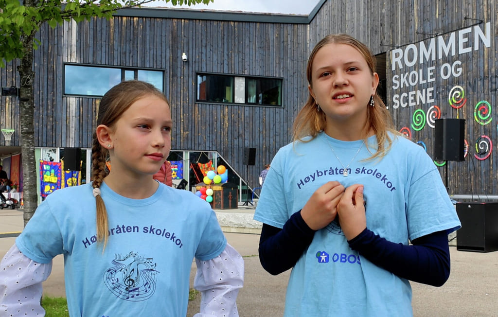 To unge jenter i lyseblå t-skjorter står utendørs i nærheten av en bygning med skilt bak seg.