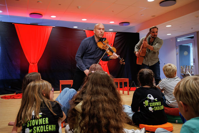 To musikere som spiller fiolin og gitar opptrer for barn som sitter på gulvet på Stovner Skolekor-arrangementet.