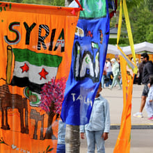 Fargerike håndmalte bannere som representerer Syria henger mellom trær på en utendørs kulturfestival, med folk som går i nærheten.