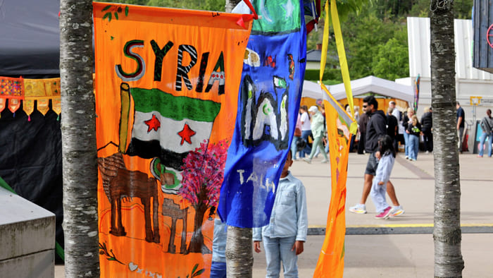 Fargerike håndmalte bannere som representerer Syria henger mellom trær på en utendørs kulturfestival, med folk som går i nærheten.
