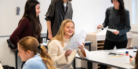 Elever og lærere i klasserommet, fokusert på jente som ser på noen tegninger. Andre engasjerer seg i ulike aktiviteter rundt bordet.