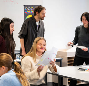 Elever og lærere i klasserommet, fokusert på jente som ser på noen tegninger. Andre engasjerer seg i ulike aktiviteter rundt bordet.