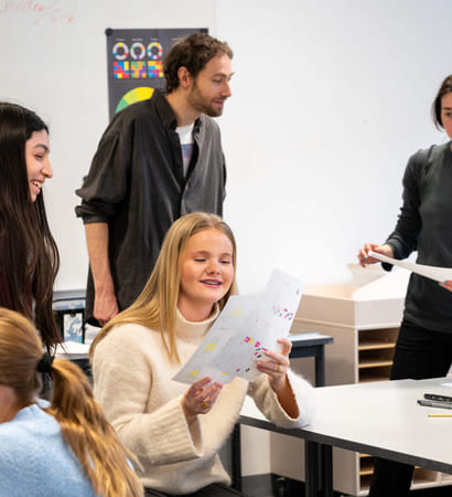 Elever og lærere i klasserommet, fokusert på jente som ser på noen tegninger. Andre engasjerer seg i ulike aktiviteter rundt bordet.