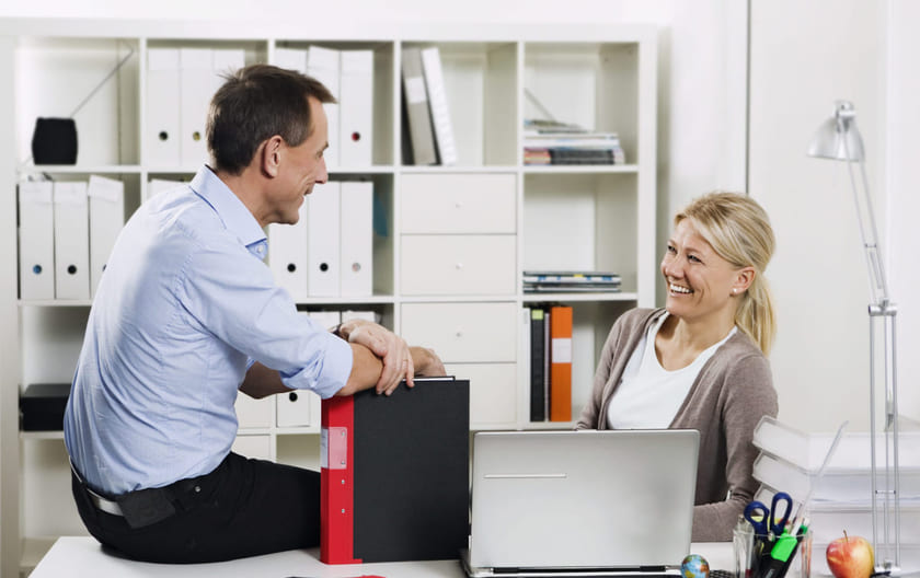 Hyggelig blid leder sitter på skrivebordet til en medarbeider. Begge smiler.