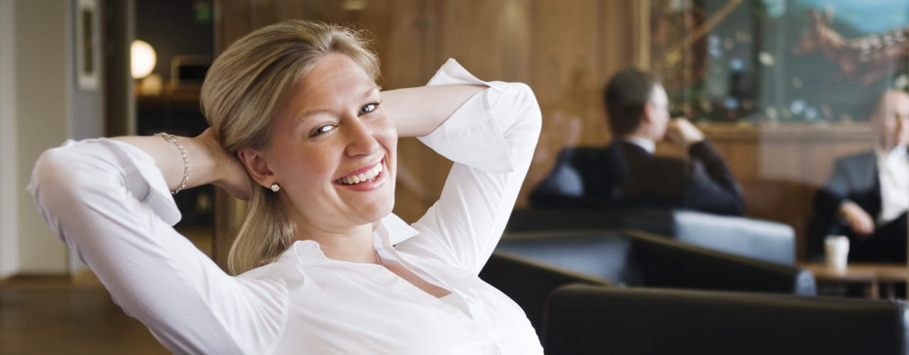 Kvinne i hvit skjorte sitter tilbakelent og ser fornøyd ut. I bakgrunnen anes et par menn.