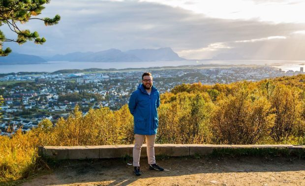Ung mann i lang, blå jakke står foran et høstlandskap.