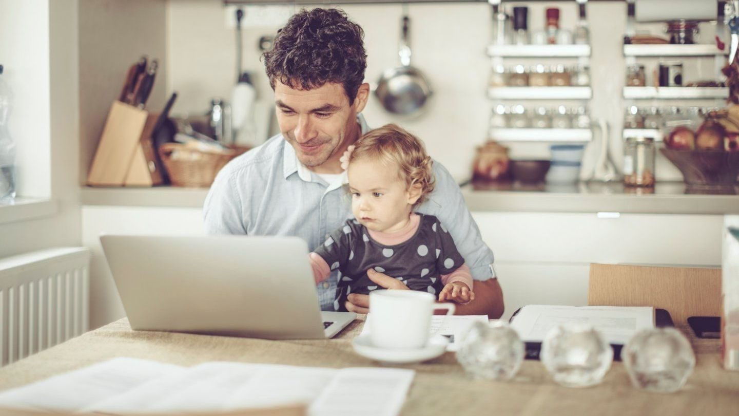 Man sitter på hjemmkontor foran pc med baby i fanget