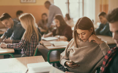 Ungdom sitter i klasserom med mange elever og ser på telefonen. Bilde.