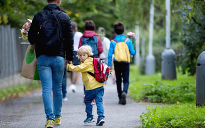 Et barn med sekk på ryggen holder en voksen i hånda mens de går bortover et fortau. Bilde.