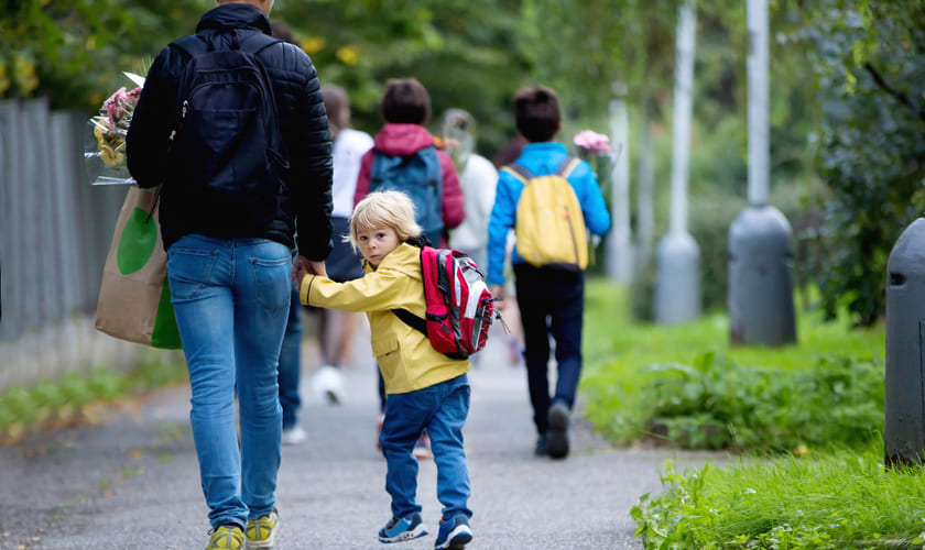 Et barn med sekk på ryggen holder en voksen i hånda mens de går bortover et fortau. Bilde.