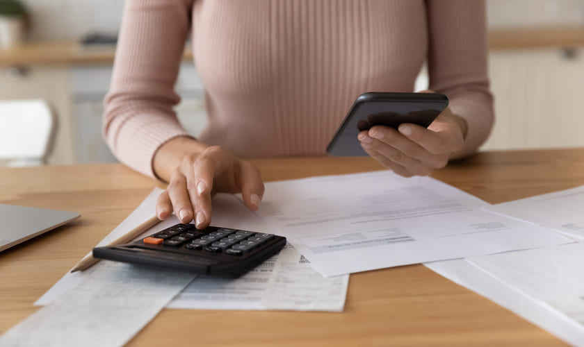 Dame sitter med kalkulator og mobil og regninger ved et bord. Bilde.