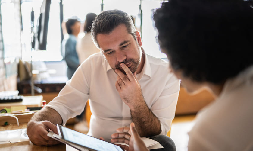 Mann sitter ved bord og ser på et nettbrett med en kollega. Bilde.