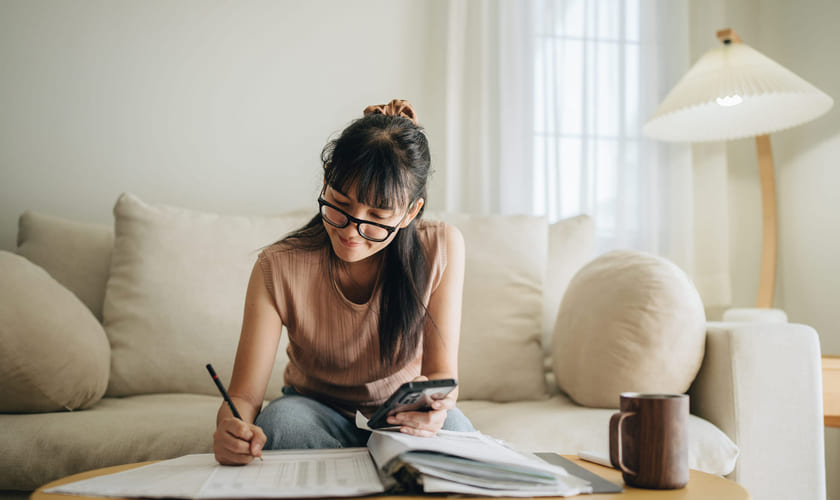 En ung kvinne sitter ved stuebordet med papirer og en kalkulator. Bilde.