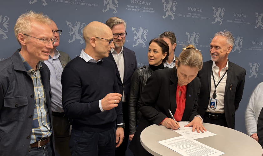 Mennesker står rundt et bord hvor Akademiker-lederen signerer den nye IA-avtalen. Bilde.
