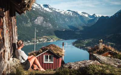 Kvinne sitterved veggen på en seter og ser ut over en fjord. Bilde.