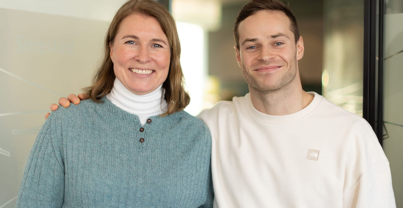 en kvinne med brunt hår og blå genser og en mann med brunt hår og hvit genser står ved siden av hverandre og smiler inn i kameraet i et kontorlandskap.