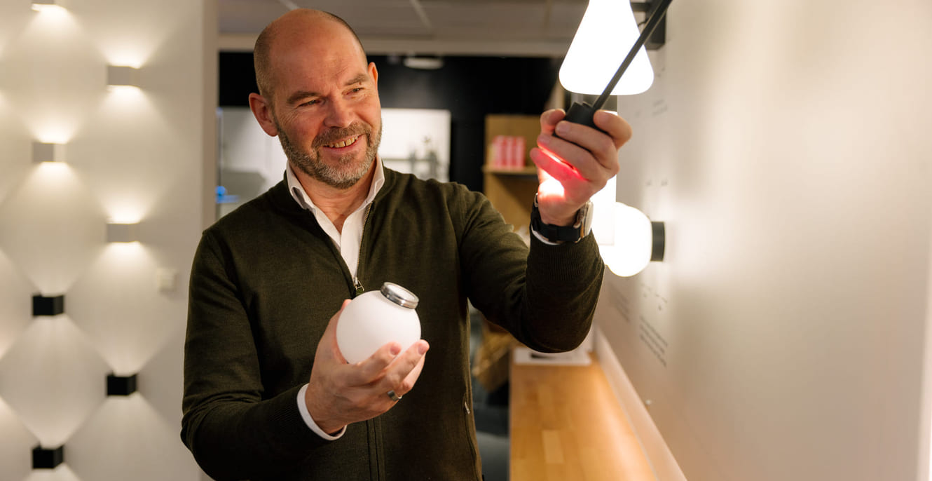 En skallet mann med grønn genser og hvit skjorte skrur av pæren på en lampe i et showroom.