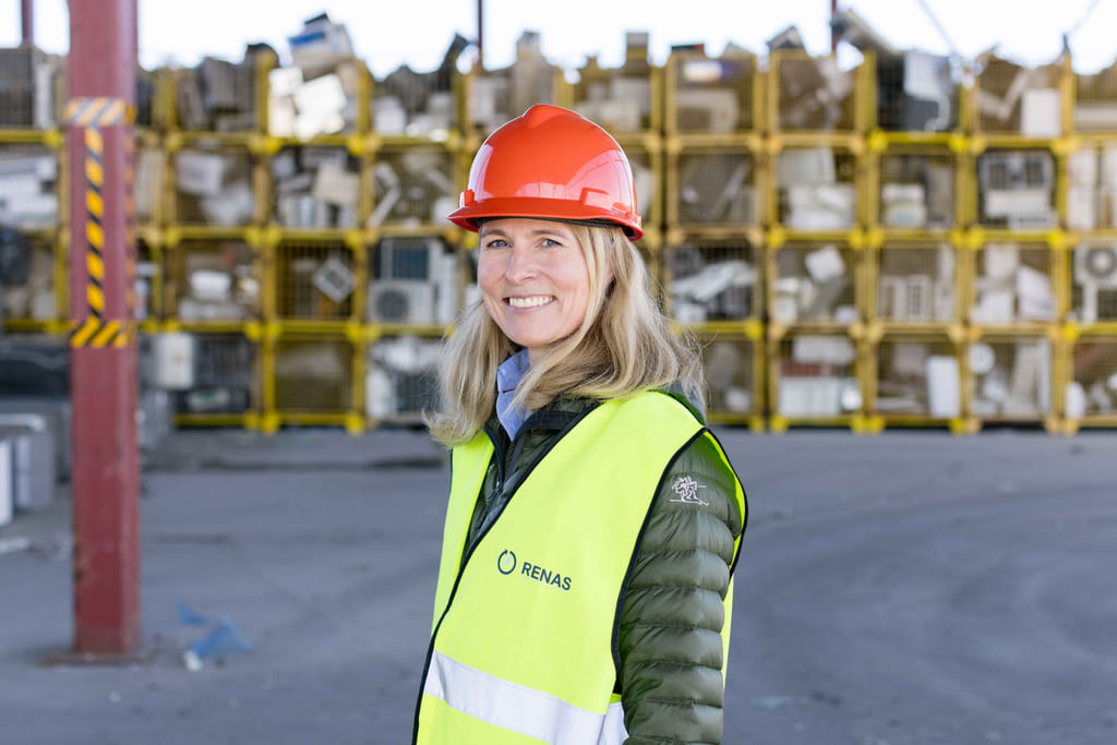 En kvinne iført hjelm og varselvest står foran stablede kasser fylt med elektronisk avfall på et gjenvinningsanlegg.