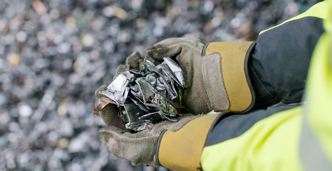 En person iført hansker og varseljakke holder en håndfull knust glass eller metallsplinter over en uskarp bakgrunn av lignende materiale.
