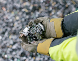 En person iført hansker og varseljakke holder en håndfull knust glass eller metallsplinter over en uskarp bakgrunn av lignende materiale.