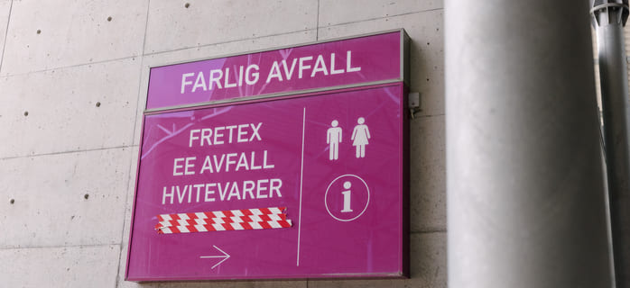 Et lilla skilt på en betongvegg viser vei på norsk, med ikoner for informasjon, og en pil som peker til høyre.