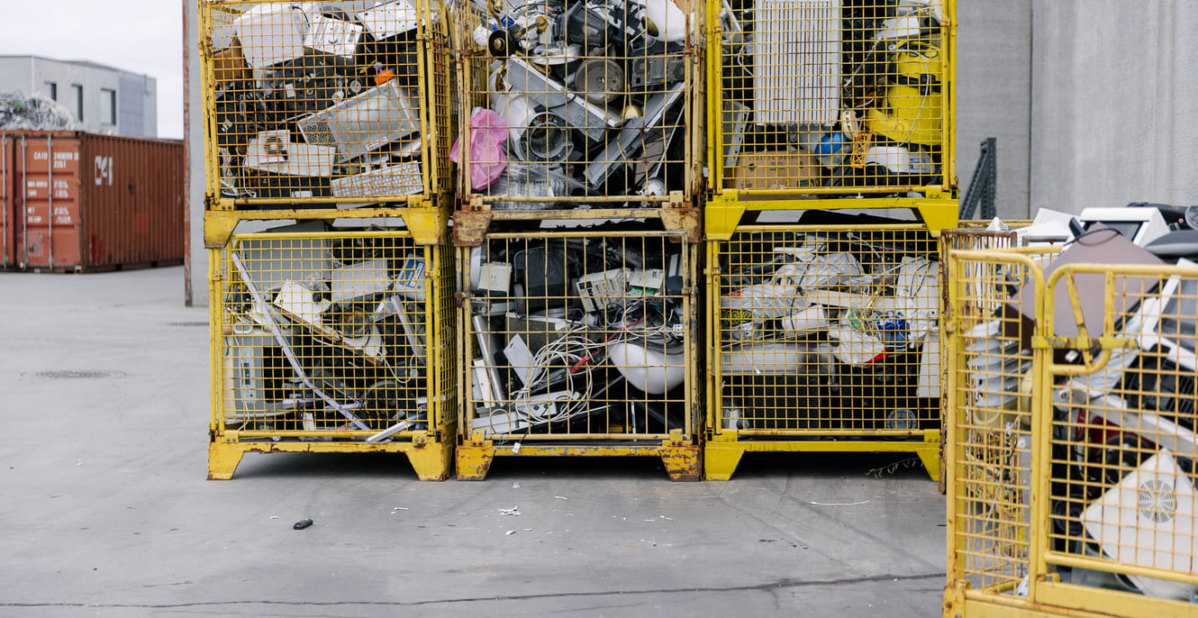 Seks gule metallbur fylt med diverse kasserte elektroniske apparater og avfall ligger stablet utendørs på en betongflate i nærheten av en industribygning.