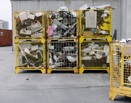 Seks gule metallbur fylt med diverse kasserte elektroniske apparater og avfall ligger stablet utendørs på en betongflate i nærheten av en industribygning.