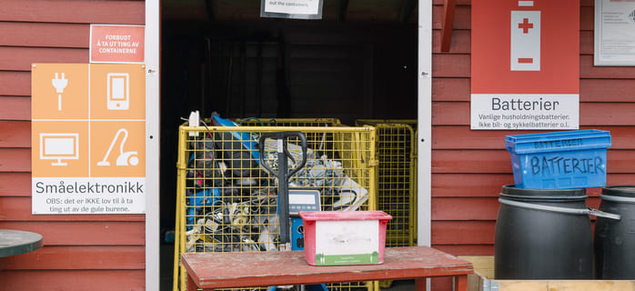 Et gjenvinningsstasjonsområde med beholdere for småelektronikk og batterier, et bur fylt med elektronisk avfall og instruksjonsskilt på norsk på en rød trebygning.