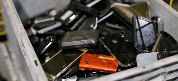 En stor plastbøtte fylt med gamle smarttelefoner og mobile enheter, inkludert ulike modeller og en oransje telefon, klargjort for resirkulering eller avhending.