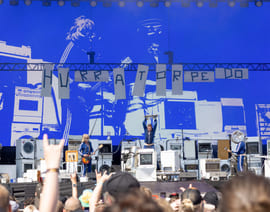 Et band opptrer på en utendørsscene med vaskemaskiner som kulisser, en stor blå skjerm i bakgrunnen og et publikum som ser på i forgrunnen.