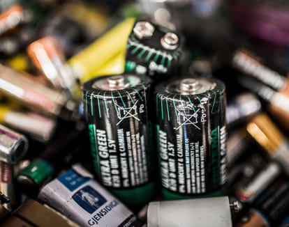 Nærbilde av ulike brukte batterier, med fokus på flere grønne og svarte AA-batterier med resirkulerings- og avhendingssymboler.