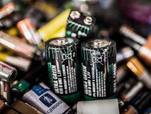 Nærbilde av ulike brukte batterier, med fokus på flere grønne og svarte AA-batterier med resirkulerings- og avhendingssymboler.