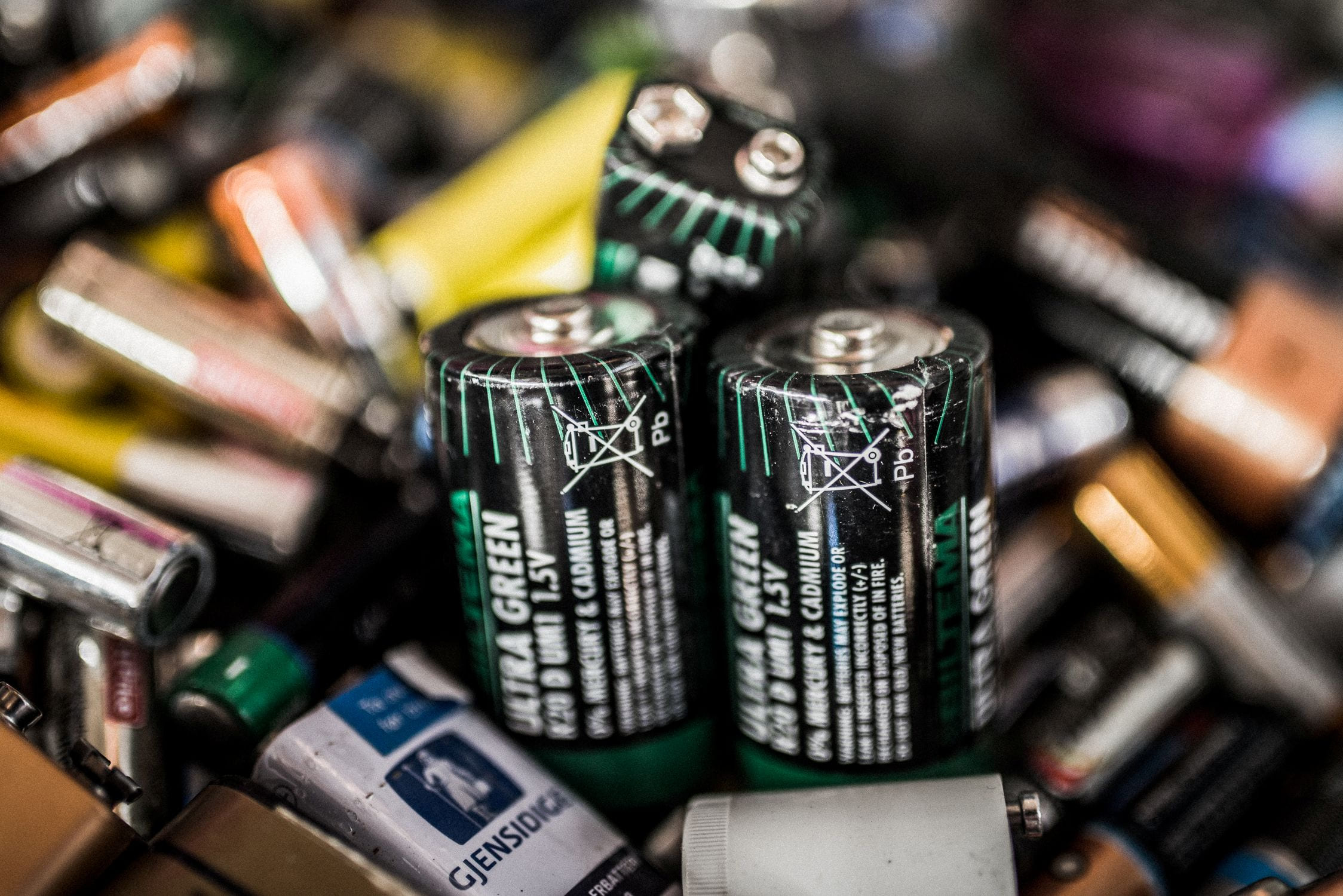 Nærbilde av ulike brukte batterier, med fokus på flere grønne og svarte AA-batterier med resirkulerings- og avhendingssymboler.