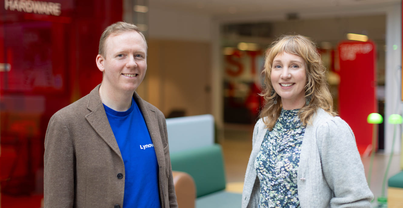 Mann i blazer og blå t-skjorte står ved siden av kvinne i cardigan og blomstrete topp i et kontorlandskap.