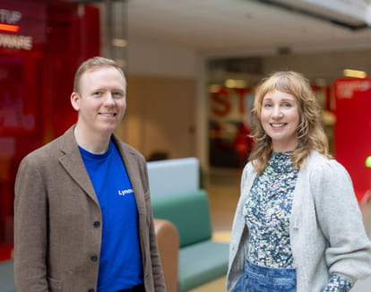 Mann i blazer og blå t-skjorte står ved siden av kvinne i cardigan og blomstrete topp i et kontorlandskap.