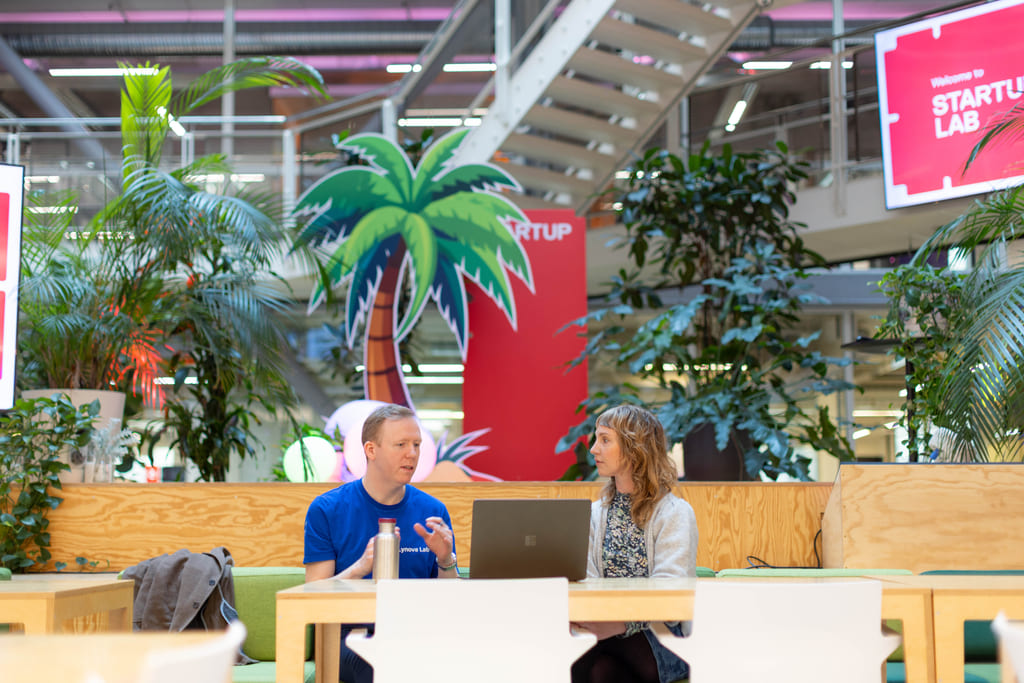 Bilde tatt langt fra, av en mann i blå t-skjorte og en kvinne i grå cardigan og blomstrete topp som sitter i et stort og åpent kontorlandskap, med en laptop foran seg.