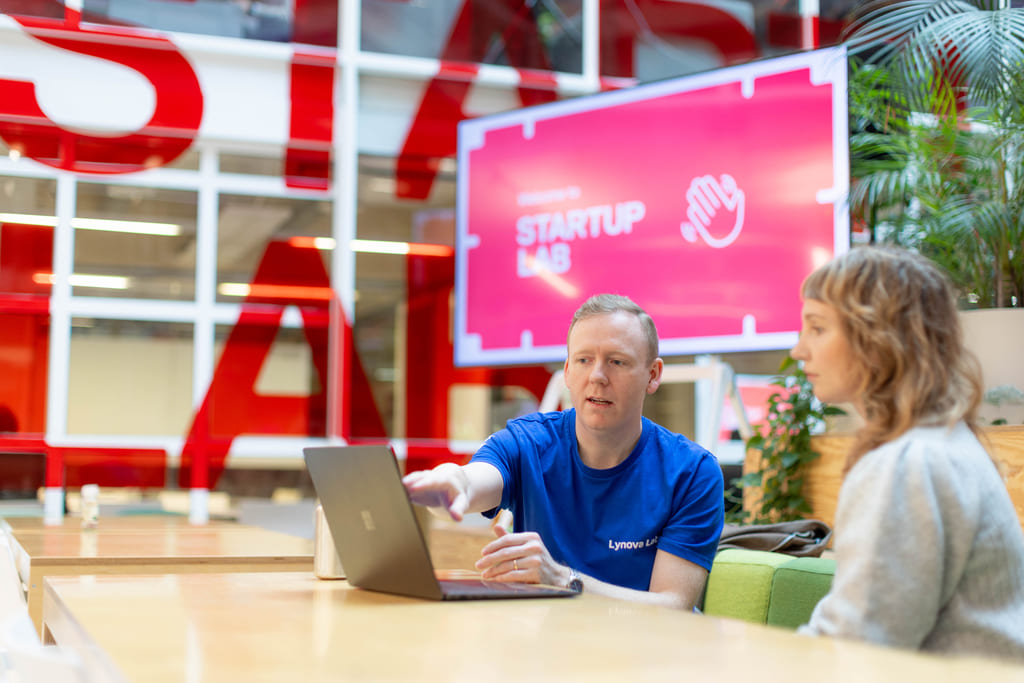 Mann i blå t-skjorte viser kvinne med grå cardigan noe på en laptop i et moderne kontorlandskap.