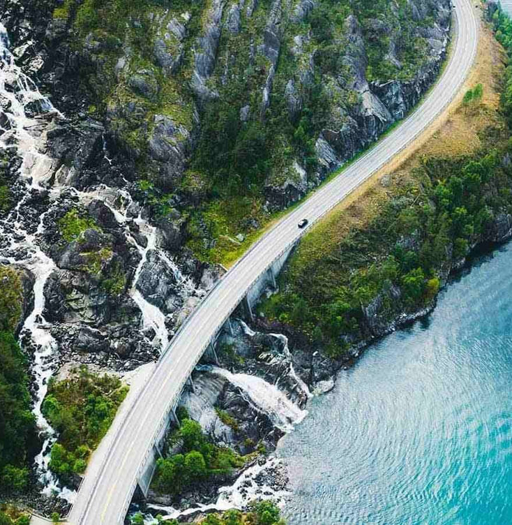 Elbil på bilvei sett ovenfra i grønt naturlandskap med fjord og fjell.