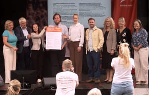 Foto fra overlevering på Kanlplasscenen i Arendal, av fylkesordfører, unge deltakere, og andre involverte